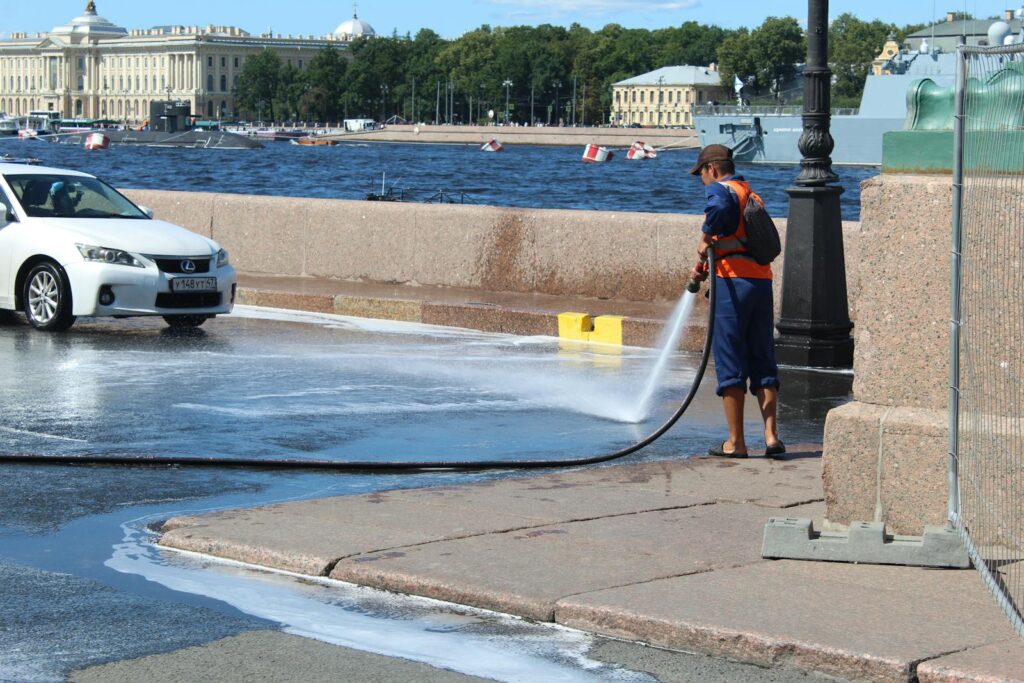 A street cleaner uses a hose to wash a sidewalk near the river, with a cityscape in the background.